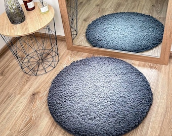 May include: A round, gray and white ombre rug with a fluffy texture. The rug is on a wooden floor.