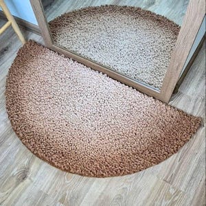 May include: A semi-circular rug with a colour gradient, fading from light beige to a darker brown. The rug has a soft, textured appearance and is positioned on a wooden floor. A mirror reflects the rug, displaying its shape and colour.