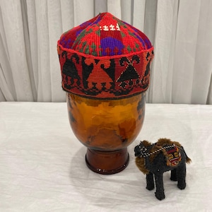 May include: A colorful, patterned cap with red, green, purple, and black designs. The cap sits atop a clear, amber-colored glass vase. A small, stuffed camel with a saddle is next to the vase.
