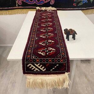 May include: A long, rectangular runner rug with a burgundy background and geometric patterns in red, white, and black. The rug has a cream-colored fringe at both ends. A small, black camel figurine sits on the table.