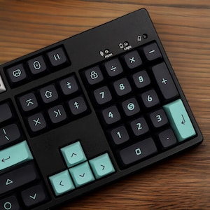 Puede incluir: Un teclado mecánico negro con teclas turquesas y gris oscuro. El teclado tiene un teclado numérico y teclas de función. Las teclas están etiquetadas con símbolos y números. El teclado está sobre una superficie de madera.