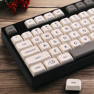 Puede incluir: Un teclado blanco y negro con teclas blancas y letras negras. Las teclas tienen varios símbolos y letras, incluyendo ESC, TAB, CAPSLOCK, SHIFT, CTRL, ALT y WIN. El teclado está sobre una superficie de madera.