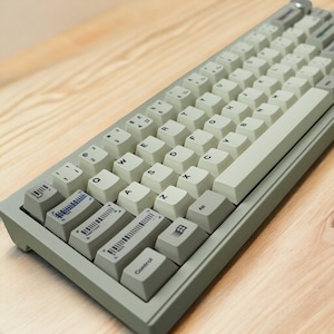 Peut inclure: Un clavier mécanique gris clair avec des touches blanches et des lettres noires. Le clavier comporte une rangée de touches de fonction en haut et une rangée de touches modificatrices en bas. Le clavier est sur une surface en bois clair.