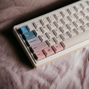 May include: A white mechanical keyboard with pastel pink, blue, and beige keycaps. The keyboard has a cream-coloured frame and is sitting on a light brown fabric surface. The keys are labelled with the standard alphabet and number keys, as well as function keys such as Tab, CapsLock, Shift, Ctrl, and Alt.