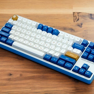 May include: A blue and white mechanical keyboard with gold keycaps. The keyboard has a standard layout with function keys, arrow keys, and a number pad. The keyboard is on a wooden surface.