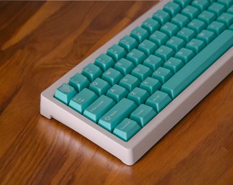 Peut inclure: Un clavier mécanique blanc et turquoise avec une barre d'espace turquoise et une touche blanche avec un petit motif. Le clavier est posé sur une surface en bois.