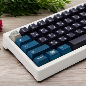 May include: A white and blue mechanical keyboard with a dark gray keycap set. The keyboard has a white frame and a dark gray bottom row. The keys are labeled with letters and numbers. The keyboard is on a wooden surface.