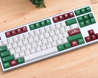 May include: A white mechanical keyboard with green and red keycaps featuring camping-themed icons. The keycaps have icons such as a tent, a mountain, a campfire, and a tree. The keyboard has a white space bar and a silver frame.