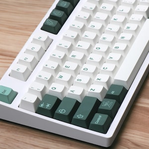 May include: White mechanical keyboard with green keycaps. The keyboard has a white case and a white space bar. The keycaps are green and have a variety of symbols and letters on them. The keyboard is on a wooden surface.