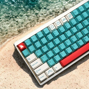 Può includere: Una tastiera meccanica bianca con tasti color turchese e una barra spaziatrice rossa. La tastiera ha un cappuccio del tasto a forma di pinna di squalo e un cappuccio del tasto a forma di ancora. La tastiera è su una superficie sabbiosa con acqua blu sullo sfondo.