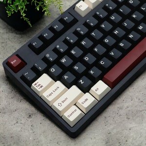 Può includere: Una tastiera meccanica nera con tasti bianchi e un tasto di accento rosso. La tastiera ha un layout standard con tasti funzione, tasti numerici e tasti alfabetici. I tasti hanno etichette di testo per ogni tasto, tra cui "Esc", "Tab", "Caps Lock", "Shift", "Control", "Code" e "Alt".