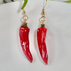 May include: A pair of red chili pepper dangle earrings with gold hooks. The chili peppers are shiny and have a realistic appearance.