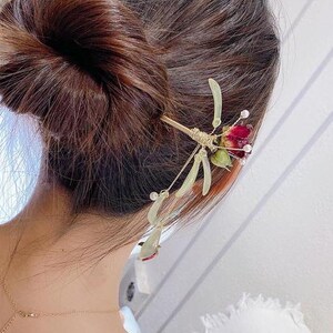 May include: A gold hair stick with a red and green flower design. The stick is decorated with small beads and has a long, thin stem.