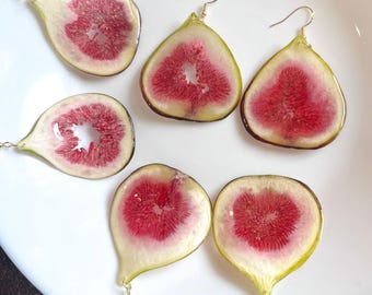 May include: A pair of earrings made from dried fig slices. The earrings are a light green color with a red center and have gold hooks.