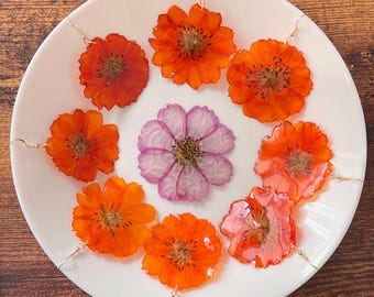 May include: A collection of handmade floral earrings and a single flower on a white plate. The earrings feature vibrant orange flowers with gold-colored hooks. A single pink flower is in the center. The plate sits on a wooden surface.