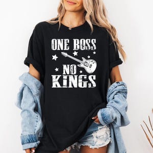 Puede incluir: Camiseta negra con el texto blanco "ONE BOSS NO KINGS", un gráfico de guitarra eléctrica blanca y estrellas. La camiseta es de estilo casual, adecuada para el uso diario.