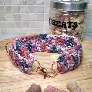 Può includere: Un collare per cani in crochet rosa, blu e marrone con una fibbia in metallo dorato rosa. Il collare è appoggiato su una superficie di legno con bocconcini per cani nelle vicinanze.