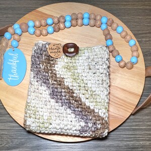 May include: A crocheted phone case with a brown and white striped pattern. The case has a wooden button and a tag that says "Hand Made". The case is on a wooden tray with a blue and brown beaded necklace.