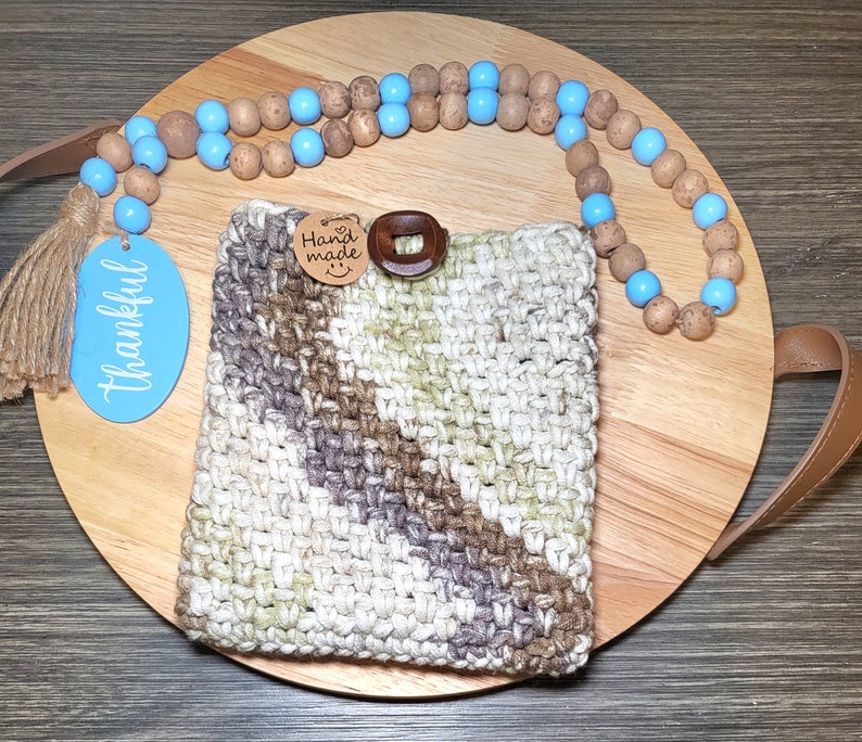May include: A crocheted phone case with a brown and white striped pattern. The case has a wooden button and a tag that says "Hand Made". The case is on a wooden tray with a blue and brown beaded necklace.