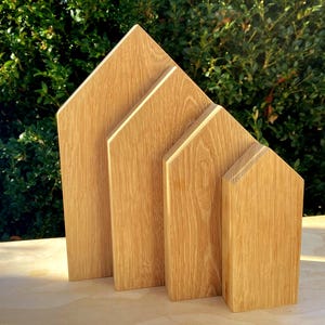 Könnte beinhalten: Vier holzförmige Hausblöcke unterschiedlicher Größe, in einer Reihe angeordnet. Die Blöcke bestehen aus hellem Holz mit sichtbarer Maserung und stehen auf einer hellen Oberfläche. Der Hintergrund zeigt grünes Laub.