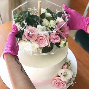 Könnte beinhalten: Eine zweistöckige weiße Torte, die mit rosa und weißen Blumen verziert ist. Die Torte ist mit einer durchsichtigen sechseckigen Box gekrönt, die mit rosa und weißen Blumen gefüllt ist.