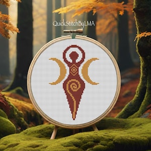 Puede incluir: Un patrón de punto de cruz de una diosa de la triple luna roja y dorada sobre un fondo blanco. El patrón está enmarcado en un aro de madera y se muestra sobre una superficie cubierta de musgo.