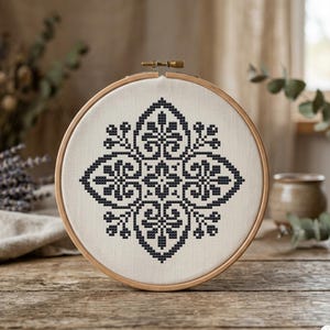 May include: A circular wooden embroidery hoop containing a piece of off-white fabric with a symmetrical black cross-stitch pattern featuring floral and geometric motifs. The design is centered on the fabric, which is held taut by the frame.