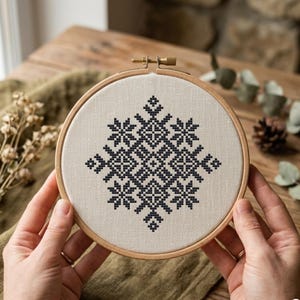 以下が含まれることがあります： 木製の刺繍枠に入った、完成したクロスステッチプロジェクト。デザインは、ベージュの生地に黒い糸で幾何学模様が施されています。木製のテーブルと緑を背景に、手で枠を持っています。