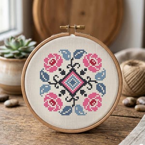 May include: A circular wooden embroidery hoop containing a cross-stitch pattern on off-white fabric. The design features a central diamond shape surrounded by four pink floral motifs and blue leaves, all outlined in black thread stitches.
