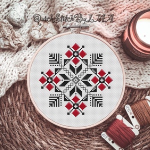 May include: A white embroidery hoop with a black and red geometric pattern. The hoop is on a woven surface with a candle and yarn.