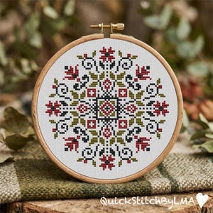 May include: A completed cross-stitch embroidery in a wooden hoop. The design features a symmetrical floral pattern in red, black, and olive green on a white background. The text "QuickStitchByLMA" is visible at the bottom.