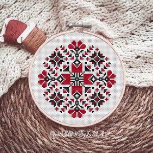 May include: A cross-stitch embroidery hoop featuring a geometric floral design in red, black, and white. The design is centered on a white fabric, held within a light pink hoop. Spools of red and brown thread are visible.