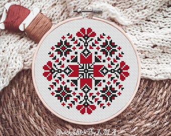 May include: A cross-stitch embroidery hoop featuring a geometric floral design in red, black, and white. The design is centered on a white fabric, held within a light pink hoop. Spools of red and brown thread are visible.