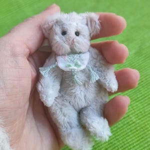 Könnte beinhalten: Ein Miniatur-Plüschteddybär mit hellgrauem, strukturiertem Fell, kleinen schwarzen Augen und einer rosa gestickten Nase. Der Bär trägt ein kleines weißes Stofflätzchen mit einem zarten Blumenmuster in Pastellgrün und Rosa, verziert mit hellgrünen Quasten.