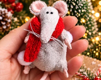 May include: A small, plush gray mouse figurine featuring a bright red knitted scarf wrapped around its neck. The mouse has pink inner ears, a long thin tail, and white whiskers. It is crafted from soft fabric with a rounded body and small limbs.