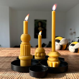 Könnte beinhalten: Drei brennende gelbe Kerzen unterschiedlicher Höhe stehen in einem schwarzen Metallkerzenhalter. Die Kerzen haben einzigartige Formen, eine mit einem gestapelten Design und eine andere mit einem abgerundeten Sockel. Die Flammen sind hell und die Szene spielt sich auf einem Holztisch ab.