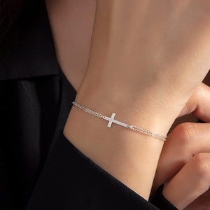 May include: A silver chain bracelet with a delicate cross charm. The bracelet is made of two thin chains and features a simple, minimalist design.