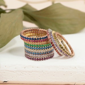 May include: A stack of colorful gemstone rings, each with a different color of stones. The rings are made of gold and are stacked on a white surface.