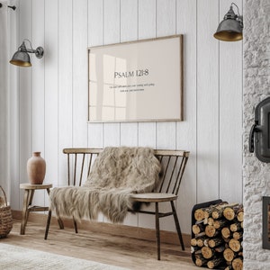 May include: A framed print with a beige background and black text that reads "PSALM 121:8" and "The Lord will watch over your coming and going both now and forevermore". The print is hanging on a white wall above a wooden bench with a furry throw blanket.