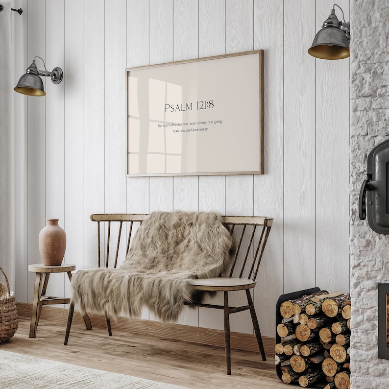 May include: A framed print with a beige background and black text that reads "PSALM 121:8" and "The Lord will watch over your coming and going both now and forevermore". The print is hanging on a white wall above a wooden bench with a furry throw blanket.