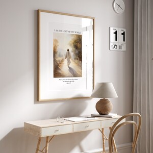 Puede incluir: Impresi&oacute;n art&iacute;stica enmarcada con el texto &laquo;I AM THE LIGHT OF THE WORLD&raquo; sobre una imagen de una figura caminando por un camino. La impresi&oacute;n est&aacute; en un marco de madera y cuelga sobre un escritorio con una l&aacute;mpara y un calendario.