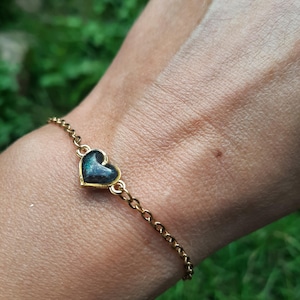 Può includere: Un braccialetto a catena color oro con un ciondolo a forma di cuore. Il ciondolo ha un aspetto marmorizzato verde scuro e nero. Il braccialetto è indossato su un polso su uno sfondo verde sfocato.