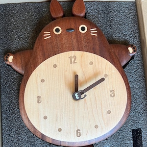 May include: A wooden wall clock shaped like a brown Totoro character with a light wood clock face. The clock face has black numbers and hands.