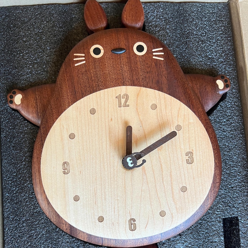 May include: A wooden wall clock shaped like a brown Totoro character with a light wood clock face. The clock face has black numbers and hands.