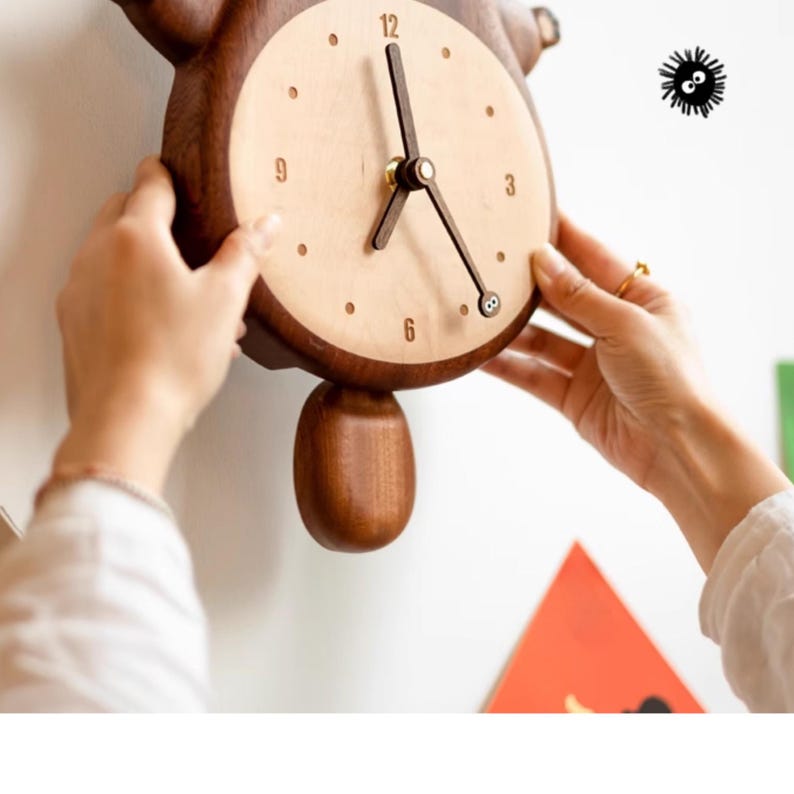 May include: A wooden wall clock shaped like a cartoon character with a round clock face and a pendulum. The clock face has black Roman numeral hour markers and black hands. The clock is being hung on a white wall.