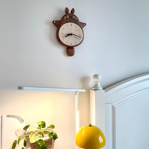 May include: A wooden wall clock shaped like a brown Totoro character with a white face and black hands. The clock is hanging on a white wall above a white bed frame with a yellow lamp.
