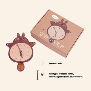 May include: A wooden wall clock shaped like the character Totoro from the movie My Neighbor Totoro. The clock has a brown wooden body and a light wood face with black hands. The clock comes with a box that has the Totoro character on it and the word "TOTORO" printed on the box. The clock also comes with two types of second hands, one with a small Totoro character and one without.
