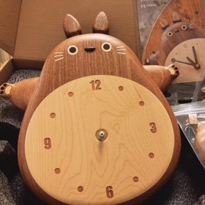 May include: A wooden wall clock shaped like a cartoon character with large ears and a round face. The clock face is light wood with dark wood numbers and hands. The character is brown with a light wood belly.