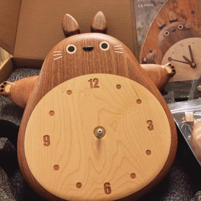 May include: A wooden wall clock shaped like a cartoon character with large ears and a round face. The clock face is light wood with dark wood numbers and hands. The character is brown with a light wood belly.