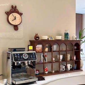 May include: A stainless steel espresso machine with a brown wooden shelf unit behind it. The shelf unit has a variety of decorative items, including a clock, a figurine, and a jar of marbles.